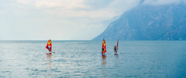 Atletas practicando windsurf en Riva del Garda, Italia, con el lago y montañas de fondo.