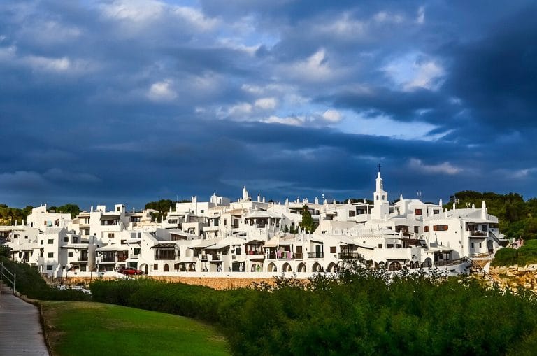 Conjunto de casas encaladas de arquitectura mediterránea en Binibeca Vell (Menorca) bajo un cielo nublado, con un pequeño paseo marítimo a la izquierda y vegetación baja en primer plano.