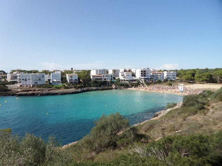 Cala Marçal con aguas turquesas cristalinas, playa de arena dorada salpicada de sombrillas y tumbonas, edificios de fachadas blancas en primera línea y vegetación mediterránea junto al acantilado bajo un cielo azul despejado.