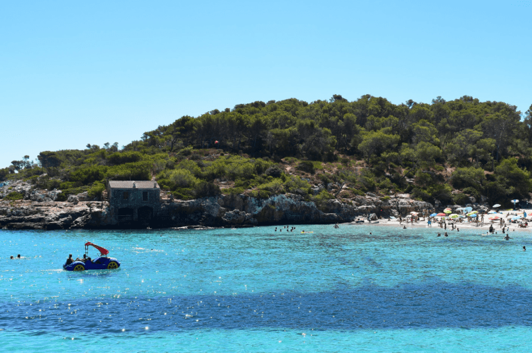 Cala Mondragó, conocida como Cala Esmeralda, muestra una pequeña bahía de aguas turquesas poco profundas rodeada de acantilados cubiertos de pinos y formaciones rocosas, con arena fina y senderos naturales que serpentean entre la vegetación mediterránea bajo un cielo claro.