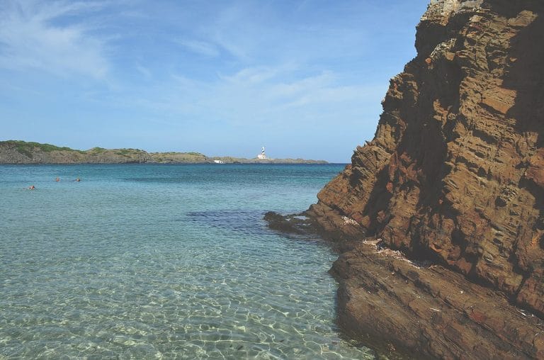 Playa de Cala Presili en Menorca, con aguas turquesas y cristalinas frente a un acantilado rojizo, formaciones rocosas sumergidas y, en el horizonte, la silueta del faro de la Illa de l’Aire bajo un cielo despejado.