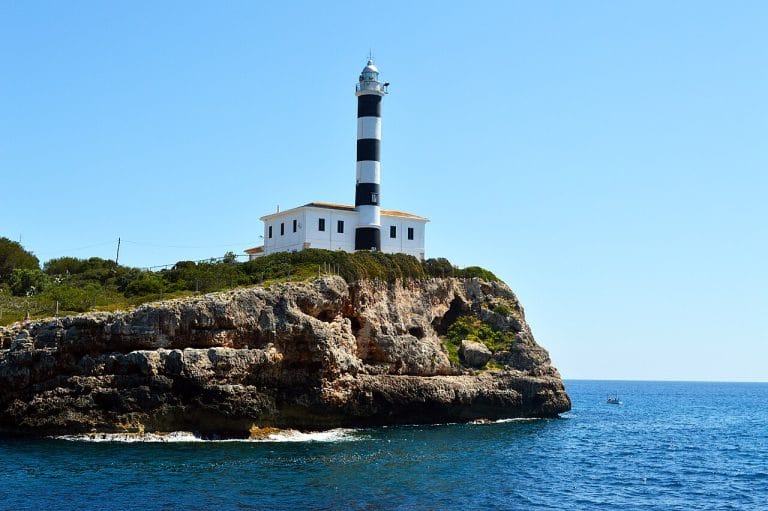 Faro de Portocolom encaramado sobre un acantilado rocoso con franjas horizontales negras y blancas, bajo un cielo despejado y con el mar azul a sus pies donde se aprecia una pequeña embarcación en la distancia.