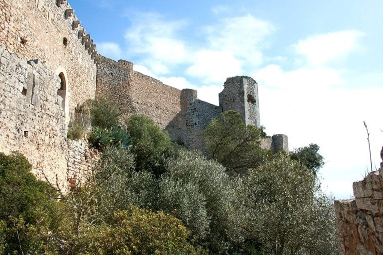 Castillo medieval en ruinas sobre un cerro cubierto de olivos y vegetación mediterránea bajo un cielo despejado.