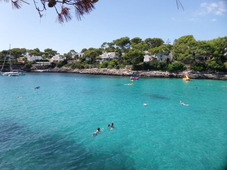 Gran Cala muestra una amplia ensenada de aguas turquesas con varias personas nadando y flotando junto a pequeñas embarcaciones, rodeada de acantilados bajos cubiertos de pinos y casas blancas bajo un cielo claro.