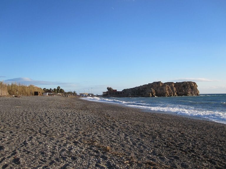 Peñón de Salobreña emergiendo junto a una playa de guijarros con olas suaves rompiendo bajo un cielo despejado.