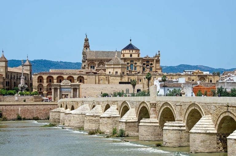 Vista del Puente Romano sobre el río Guadalquivir con arcos de piedra y la Mezquita-Catedral de Córdoba al fondo bajo un cielo azul.