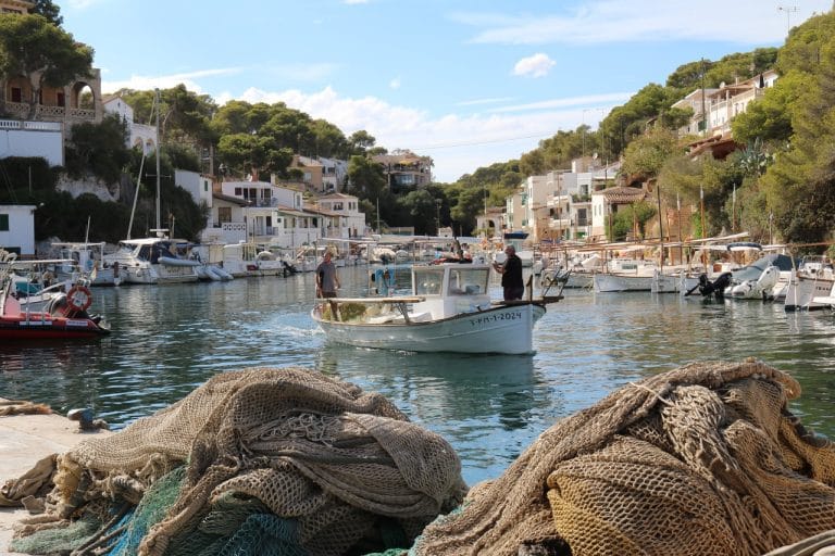 Cala Figuera muestra su pintoresco puerto pesquero con redes de pesca amontonadas en primer plano y un barco faenando en aguas turquesa rodeadas de casitas blancas y pinos mediterráneos.