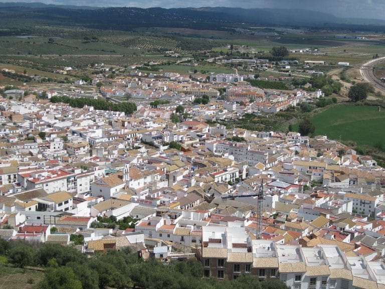 Vista aérea de Almodóvar del Río con casas blancas de tejados ocres, campos verdes y la sierra al fondo bajo un cielo nublado.
