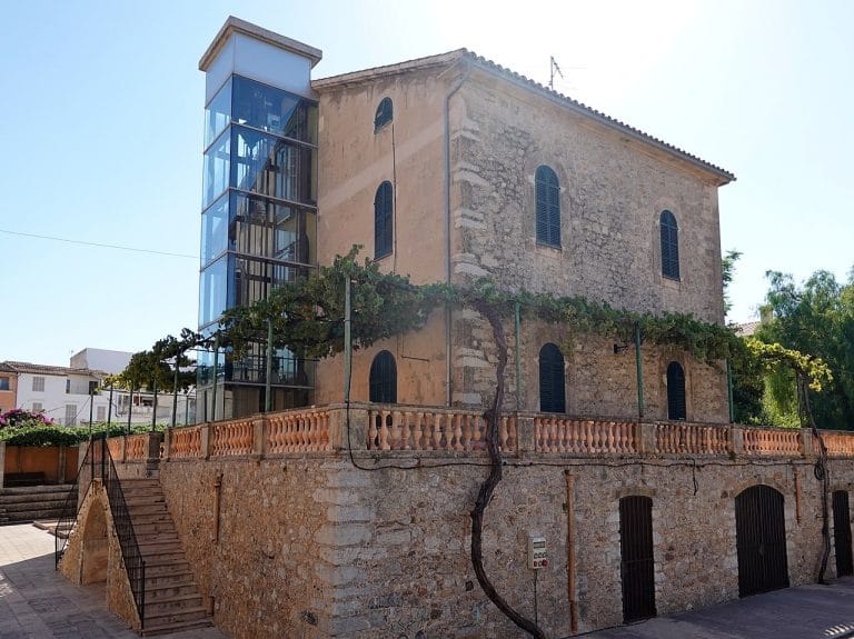 Biblioteca municipal de Artá ubicada en un edificio de piedra con un ascensor de cristal en su fachada.