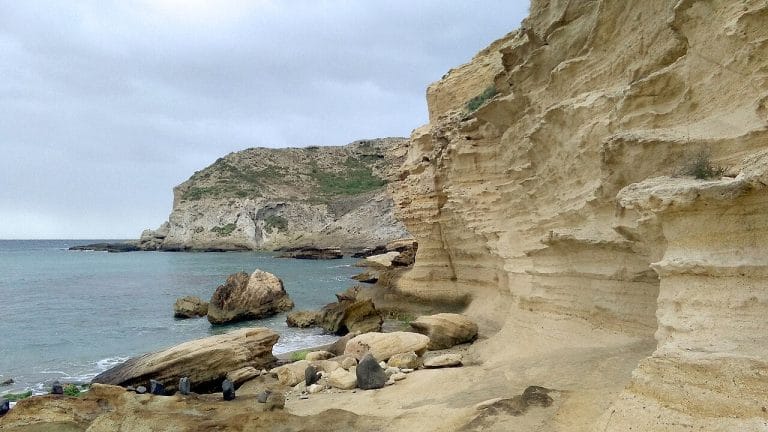 Acantilados de roca arenisca estratificada que descienden hasta una cala con rocas dispersas, aguas turquesas y cielo cubierto de nubes