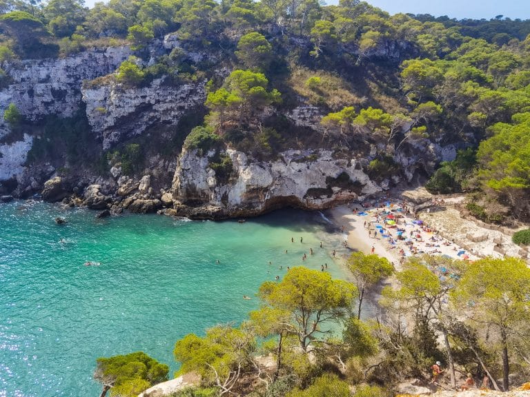 Cala Macarelleta vista desde arriba: aguas de un vibrante tono turquesa y esmeralda bañando una pequeña playa de arena rodeada de acantilados rocosos cubiertos de pinos y vegetación mediterránea.