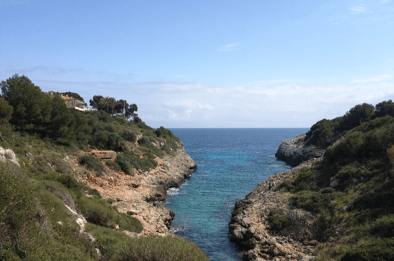 estrecho canal rocoso con aguas turquesas, acantilados cubiertos de matorral y un cielo azul con nubes dispersas.