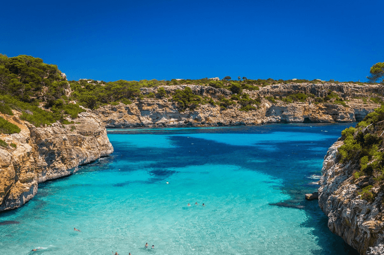 Vista aérea de Caló des Moro en Mallorca con aguas turquesa cristalinas, arena blanca y acantilados rocosos cubiertos de pinos.