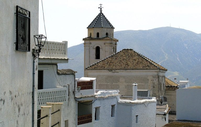 torre de iglesia y casas blancas de fachada encalada sobre una colina, con montañas de fondo bajo un cielo claro.
