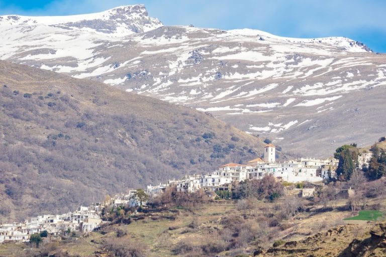 vista panorámica del pueblo blanco de capileira en la ladera de la sierra nevada con cumbres nevadas al fondo.