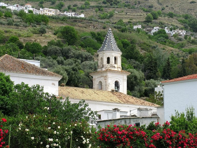 torre de iglesia con tejado de tejas y chapitel apuntado, rodeada de casas blancas y arbustos con flores, con laderas verdes al fondo.
