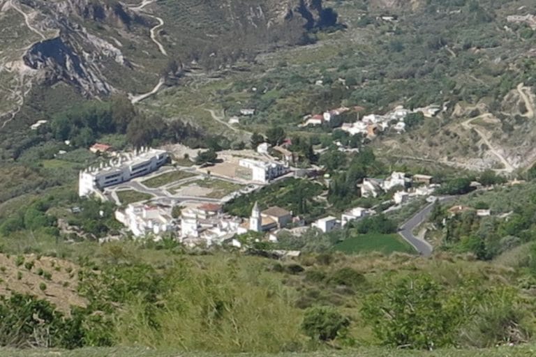 vista aérea de carataunas, con casas blancas agrupadas alrededor de una iglesia de tejado oscuro, una carretera que serpentea y colinas rocosas y terrenos agrícolas a su alrededor.