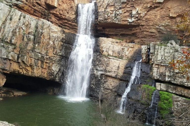 Cascada de la Cimbarra con chorros de agua que caen desde un salto elevado sobre formaciones rocosas hacia una poza rodeada de vegetación y paredes de roca.