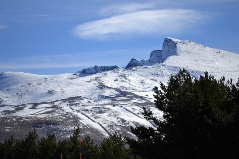 Montaña nevada en Sierra Nevada con pinos en primer plano y un cielo azul con nubes.