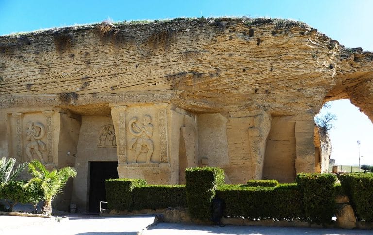 Fachada rocosa con relieves esculpidos de figuras mitológicas en el coto de las canteras bajo un cielo despejado.