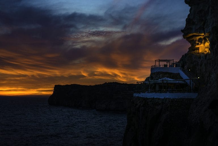 Atardecer sobre el mar desde la cova d’en xoroi, con la silueta del acantilado y la terraza iluminada donde varias personas contemplan el cielo anaranjado y púrpura.