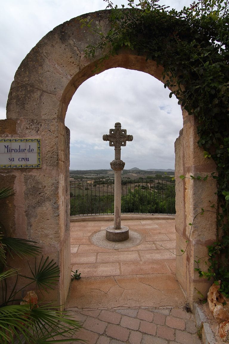 Cruz frente a la Iglesia de Nuestra Señora de Atocha en Ariany, Mallorca, con pedestal de piedra y cielo azul