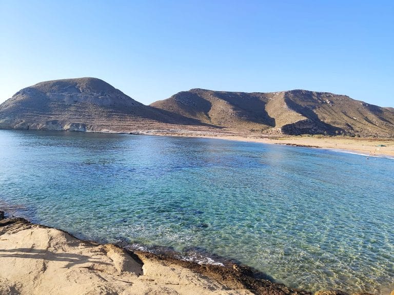 Vista panorámica de El Playazo de Rodalquilar en Cabo de Gata, con aguas cristalinas color turquesa en primer plano, arena dorada y suaves colinas áridas al fondo bajo un cielo azul despejado.
