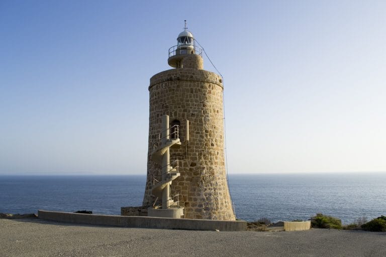 torre de faro de piedra coronada por la linterna y escalera exterior en espiral, erguida sobre un promontorio rocoso junto al mar bajo un cielo azul claro.