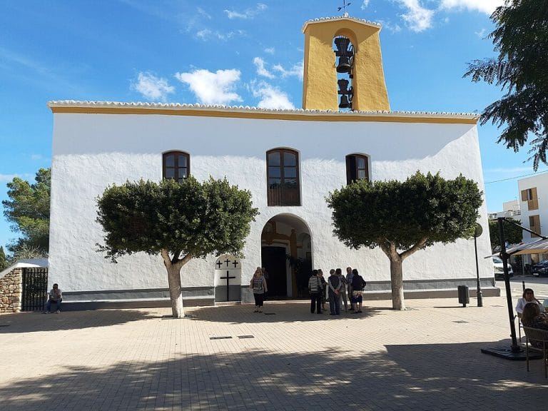 Iglesia de Santa Gertrudis en Ibiza con fachada blanca y campanario amarillo, flanqueada por dos naranjos y un grupo de personas conversando en la plaza.
