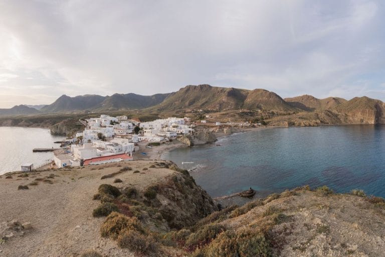 panorámica del pueblo blanco de Isleta del Moro asomado al mar en Cabo de Gata, con casas tradicionales, calas rocosas y montañas áridas al fondo bajo un cielo parcialmente nublado.