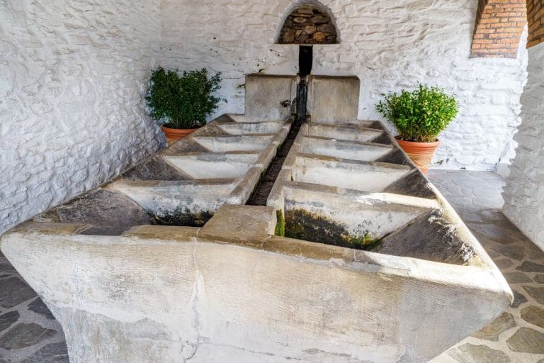interior de un antiguo lavadero público con pilas de piedra tallada, suelo empedrado y muros encalados junto a macetas.