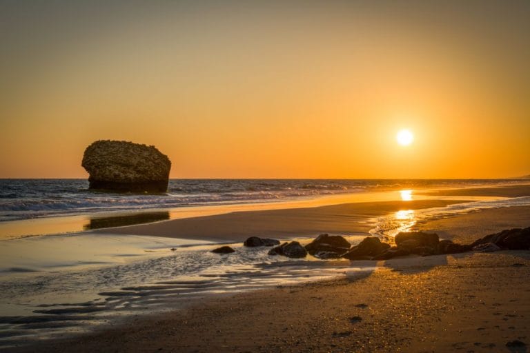 atardecer sobre una playa arenosa con reflejos dorados, un gran bloque rocoso semisumergido en el mar y el sol junto al horizonte sobre aguas tranquilas.