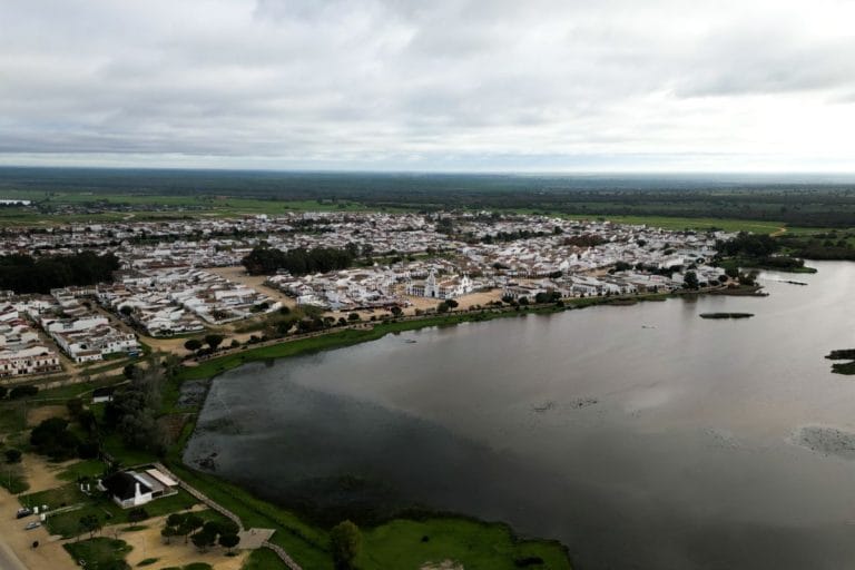 vista aérea de un pueblo de casas blancas junto a una amplia lámina de agua y campos verdes bajo un cielo nublado.