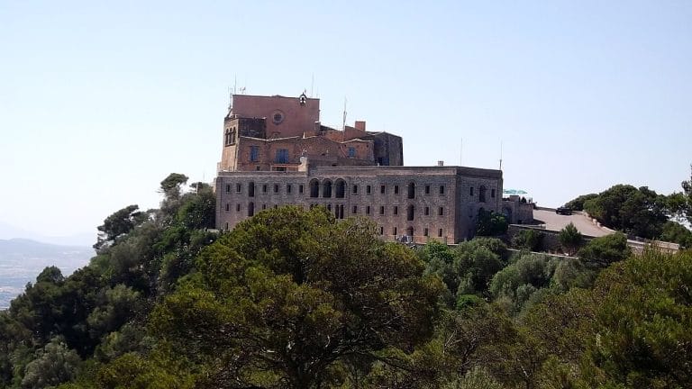 Vista frontal del monasterio de san salvador en artá, encaramado en la cima de un cerro rodeado de pinos.