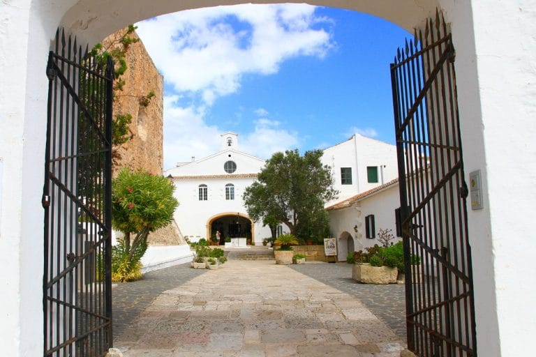 Vista del santuario de monte toro en menorca, con patio adoquinado, arcos blancos y cielo azul nublado vistos a través de una reja de hierro abierta.
