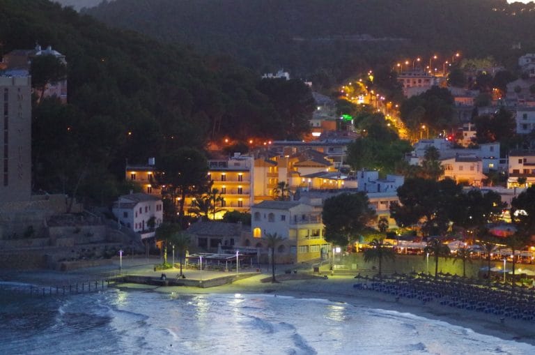 Nocturna vista de Peguera en Mallorca con luces urbanas reflejadas en las calles y siluetas de formaciones rocosas junto al mar bajo un cielo oscuro.