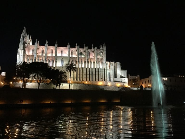 Paisaje urbano nocturno en Palma de Mallorca con la Catedral de Santa María iluminada, reflejada en una fuente, rodeada de edificios históricos.