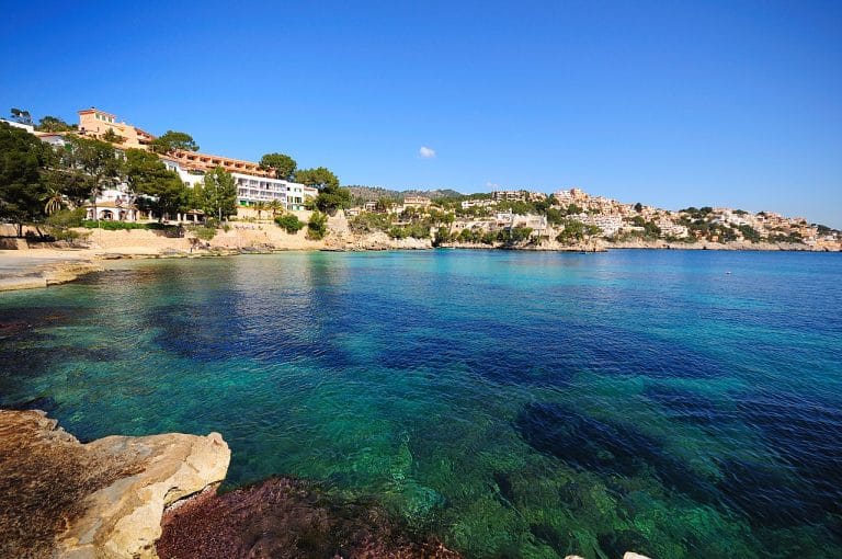 Vista de la costa de Fornells en Menorca, con aguas cristalinas color turquesa, rocas y acantilados en primer plano, casas blancas y vegetación mediterránea distribuidas en la ladera y un cielo azul claro de fondo.