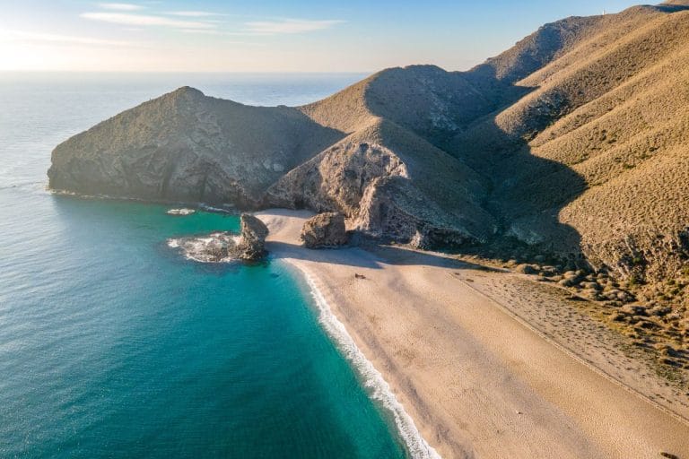 vista aérea de la playa de los Muertos en Cabo de Gata, con arena dorada, aguas turquesas y acantilados áridos al atardecer.