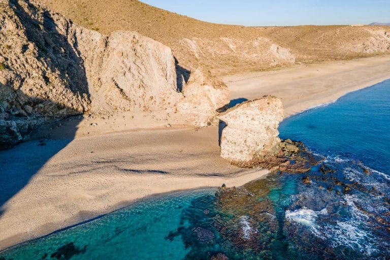 Vista aérea de una amplia playa desierta flanqueada por acantilados áridos, con una formación rocosa en forma de arco junto a la orilla y aguas turquesas del Mediterráneo.