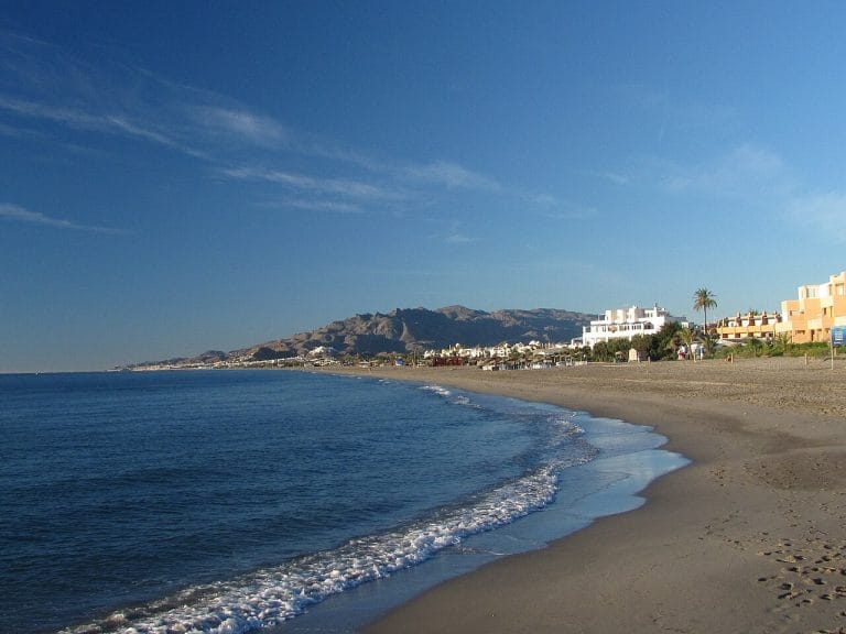 Costa de Playa El Playazo en Vera, con arena dorada y suaves olas rompiendo en la orilla, construcciones bajas y palmeras en primer plano, y montañas al fondo bajo un cielo azul.