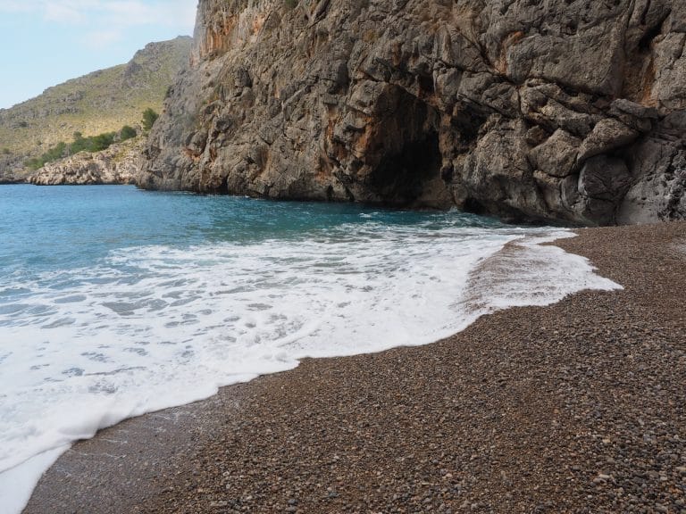 Vista de la bahía de Sa Calobra y el desfiladero del Torrent de Pareis con acantilados rocosos, aguas turquesas y arena de guijarros bajo un cielo despejado