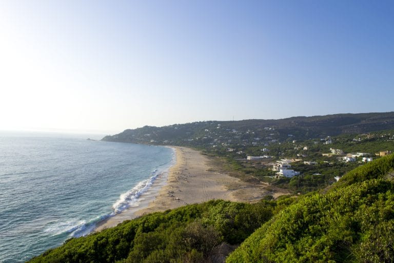 extensa franja de arena dorada bordeada por vegetación baja y casas dispersas, con olas suaves rompiendo en la orilla y un cielo despejado en el horizonte.