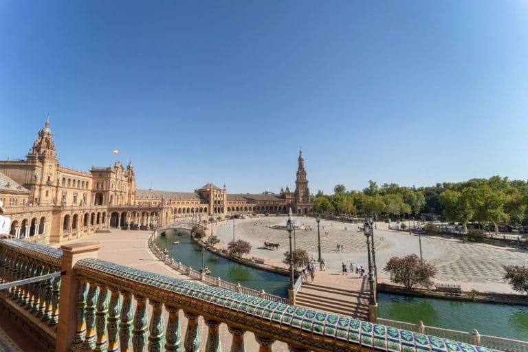 vista panorámica de la plaza de españa en sevilla con su canal en forma de semicírculo, barandillas de cerámica, la fuente central y las torres al fondo bajo un cielo azul.