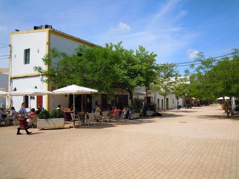Personas sentadas en la terraza de un bar en la plaza de Santa Gertrudis en Ibiza, rodeadas de árboles y edificios encalados.