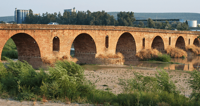 Vista lateral del puente romano de Andújar sobre el río Guadalquivir con varios arcos de ladrillo y vegetación en las orillas.