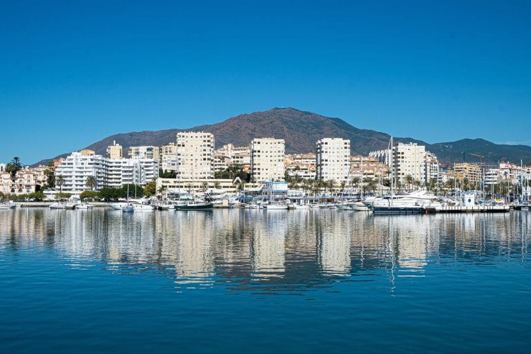 embarcaciones de recreo amarradas en el puerto deportivo de Estepona, con la línea de edificios y montañas al fondo reflejados en las tranquilas aguas azules.