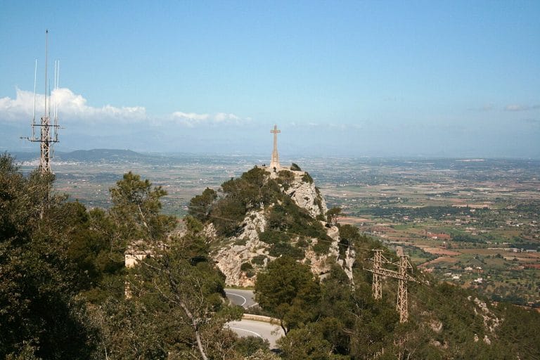Cruz monumental en lo alto del Puig de Sant Salvador con vistas panorámicas del valle bajo un cielo despejado.