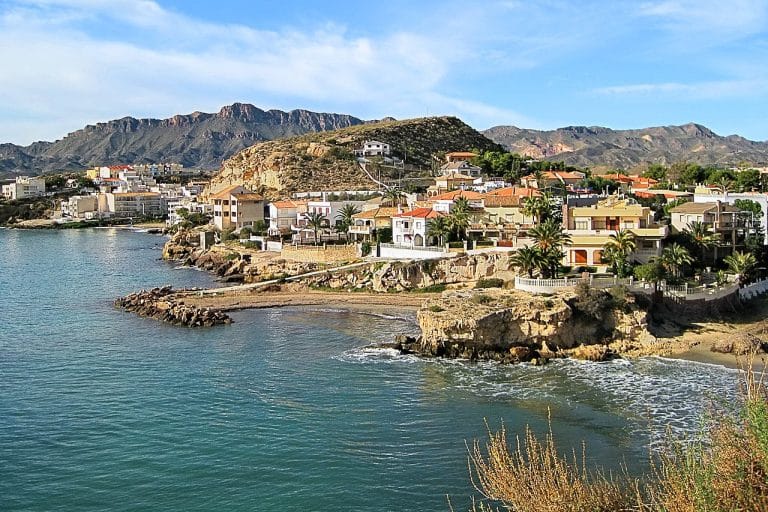 Vista panorámica de San Juan de los Terreros: casas blancas y color crema alineadas en acantilados costeros, con palmeras y vegetación, frente al mar y bajo un cielo despejado.