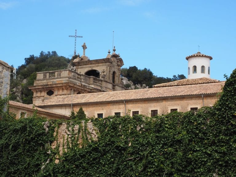 vista de la fachada del santuario de Lluc con un muro cubierto de hiedra en primer plano, la cúpula blanca y el frontón de piedra con cruces bajo un cielo azul claro.